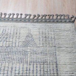 Cream shaggy rug with faint blue grid lines and braided gray tassels lies on light tan wood flooring.