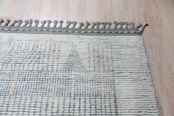 Cream shaggy rug with faint blue grid lines and braided gray tassels lies on light tan wood flooring.