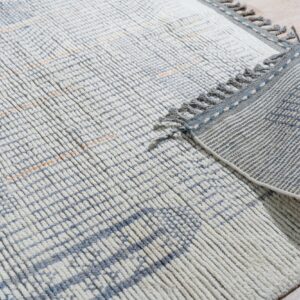 Textured white and blue rug with an orange grid pattern, fringe, and a striped backing on light flooring.