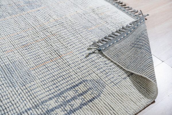 Textured white and blue rug with an orange grid pattern, fringe, and a striped backing on light flooring.