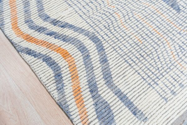Textured white rug with blue and orange intersecting patterns rests on light blonde wood flooring.