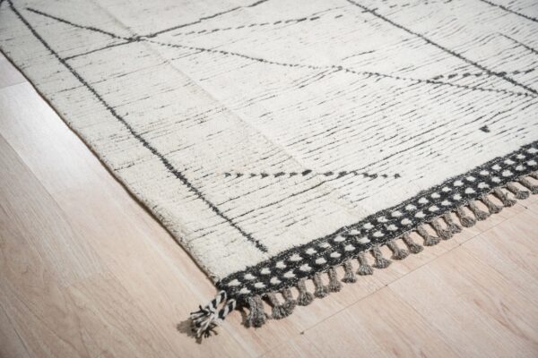 Textured white rug with dark gray geometric pattern, a dotted border, and tassels on blonde wood.
