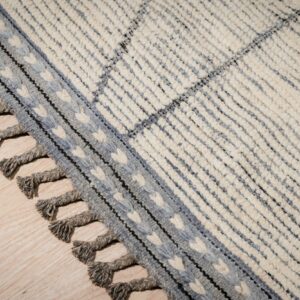 Chunky cream rug with blue-gray stripes, a patterned border, and twisted tassels on light wood flooring.