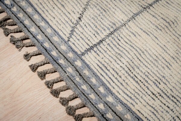 Chunky cream rug with blue-gray stripes, a patterned border, and twisted tassels on light wood flooring.