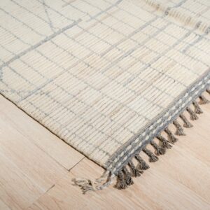 High-pile cream rug with a subtle blue diamond grid and thick grey braided fringe on light wood planks.