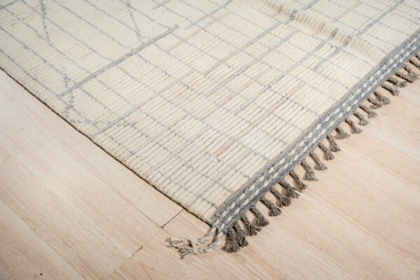 High-pile cream rug with a subtle blue diamond grid and thick grey braided fringe on light wood planks.