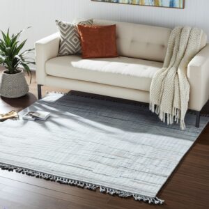 Light blue-gray rug with dark abstract lines and fringe anchors a cream sofa and potted plant on dark wood floors.
