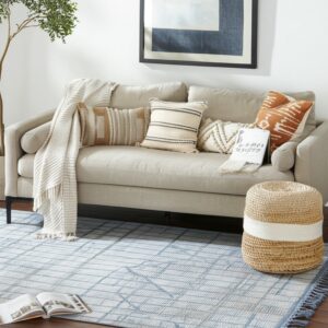 Blue and cream patterned rug with fringe on dark wood floor under a light beige sofa and wicker pouf.