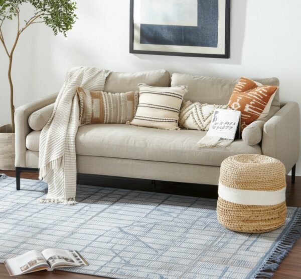 Blue and cream patterned rug with fringe on dark wood floor under a light beige sofa and wicker pouf.