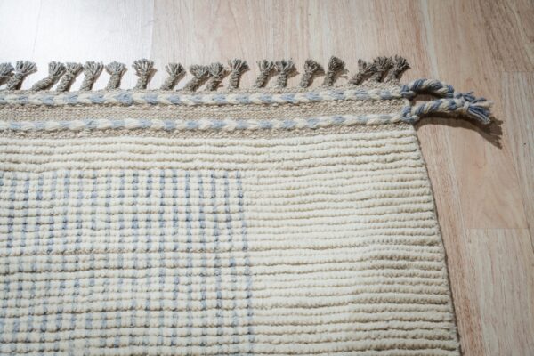 Textured cream rug with blue checkered pattern, braided borders, and gray tassels on light wood.