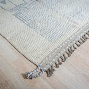 Textured cream rug features blue geometric patterns, braided fringe, and grey tassels on light wood.