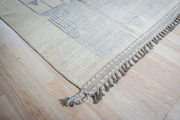 Textured cream rug features blue geometric patterns, braided fringe, and grey tassels on light wood.