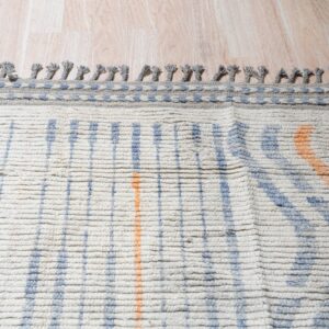 Chunky cream rug with blue and orange stripes and braided grey fringe on light wood planks.