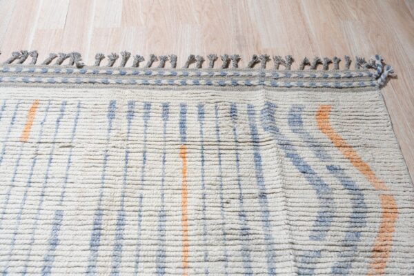 Chunky cream rug with blue and orange stripes and braided grey fringe on light wood planks.