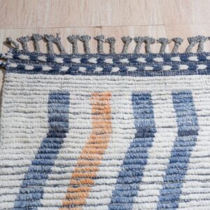 A textured cream, blue, and orange striped rug edge featuring looped gray tassels rests on light wood.