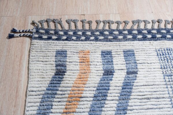 A textured cream, blue, and orange striped rug edge featuring looped gray tassels rests on light wood.
