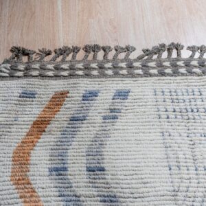 Chunky off-white rug with blue and orange geometric patterns, featuring a decorative braided gray border on wood flooring.