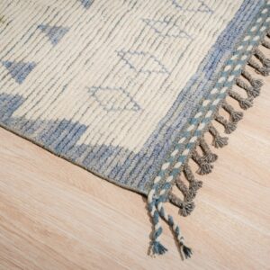 Textured cream rug with blue geometric patterns and chunky grey braided tassels on light wood flooring.