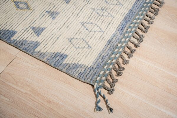 Textured cream rug with blue geometric patterns and chunky grey braided tassels on light wood flooring.