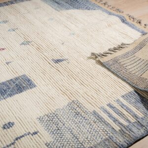 A thick, ribbed cream rug featuring abstract blue and purple patterns unrolls on light wood flooring.