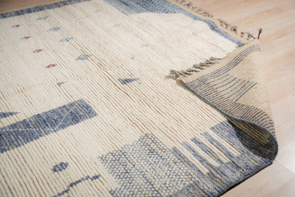 A thick, ribbed cream rug featuring abstract blue and purple patterns unrolls on light wood flooring.