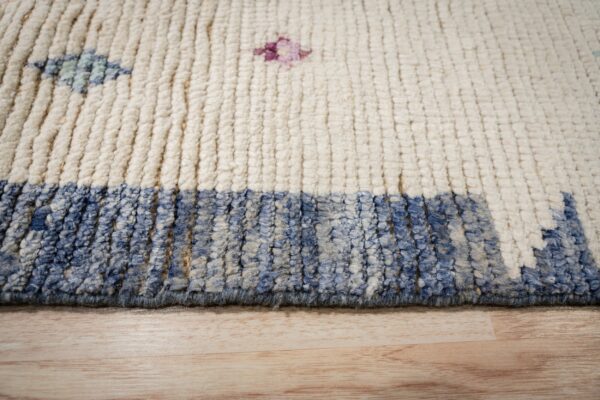 Detail of a high-pile cream rug with blue-mottled border and scattered geometric motifs on wood floor.