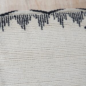 A close-up view of a cream high-pile rug with a black jagged border on light wood flooring.