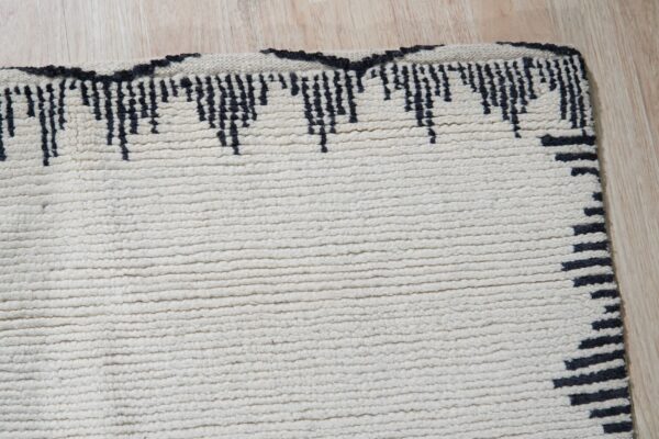 A close-up view of a cream high-pile rug with a black jagged border on light wood flooring.