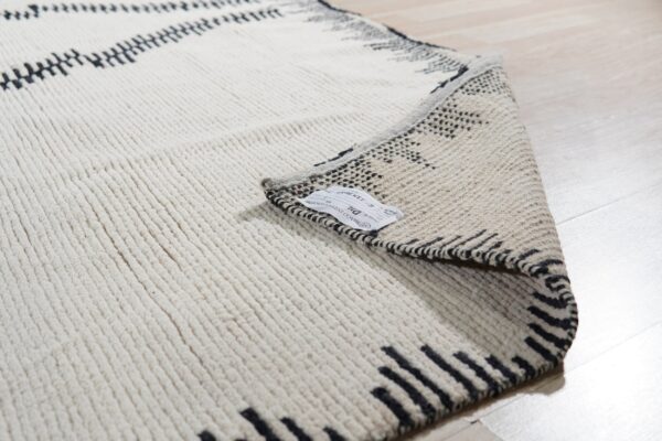 Close-up of a cream rug with black zigzag lines and a thick ribbed texture, folded over on a light wood floor.
