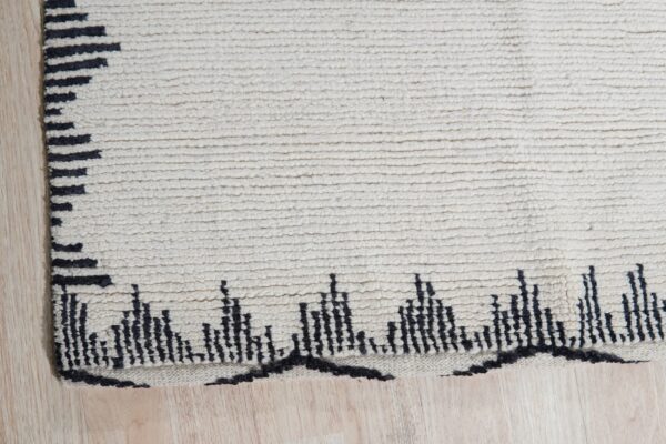 Off-white high-pile rug with a ribbed texture and black geometric patterns resting on light wood flooring.