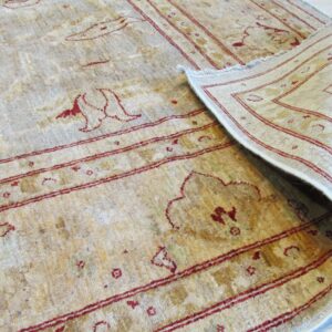 Beige rug with subtle red patterned border and motifs, curled back revealing its backing on a pale wood floor.
