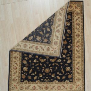 A dark blue and beige traditional patterned rug lies on light wood flooring, with a corner folded over.