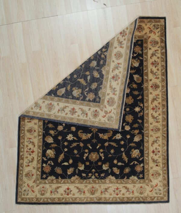 A dark blue and beige traditional patterned rug lies on light wood flooring, with a corner folded over.