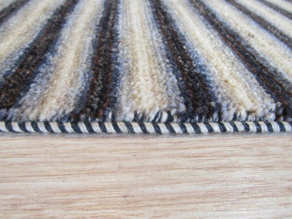 Close-up shows shaggy cream, brown, and blue striped rug pile and binding on light wood floor.