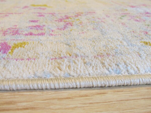 Cream low-pile rug with pink and yellow abstract pattern rests on wood flooring.