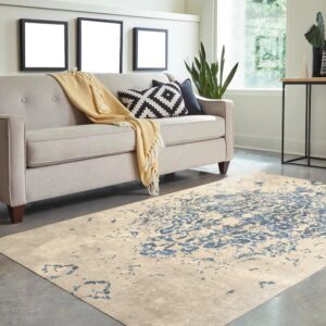 Beige rug with distressed blue pattern sits on a concrete floor beneath a light gray tufted sofa.