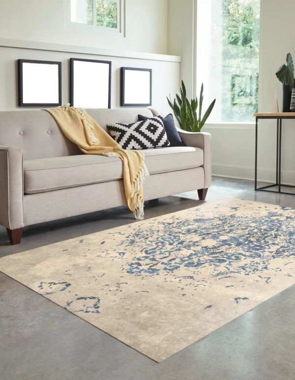 Beige rug with distressed blue pattern sits on a concrete floor beneath a light gray tufted sofa.