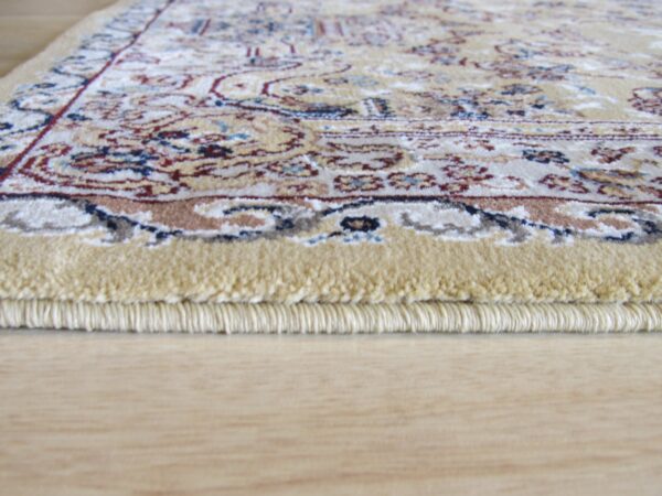 Side view of a thick, tan-edged rug showing a multicolored traditional pattern on light hardwood.