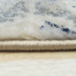 Close-up shows a thick pile rug edge with white, gray, and dark blue abstract colors over light tan flooring.