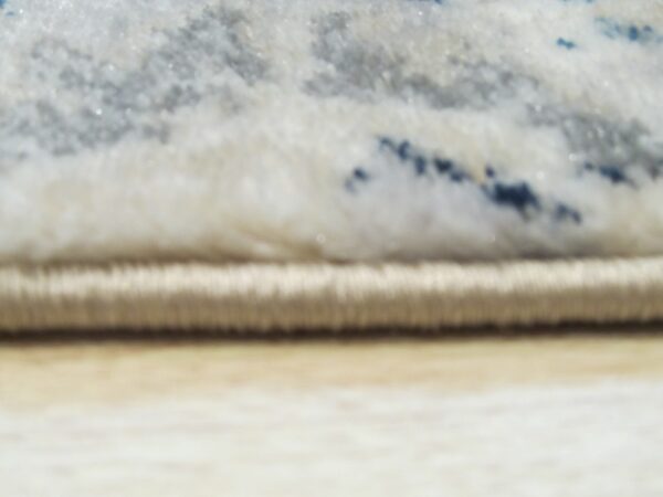 Close-up shows a thick pile rug edge with white, gray, and dark blue abstract colors over light tan flooring.