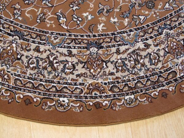 The curved edge of a brown, traditional rug with beige and blue accents lies on wood flooring.