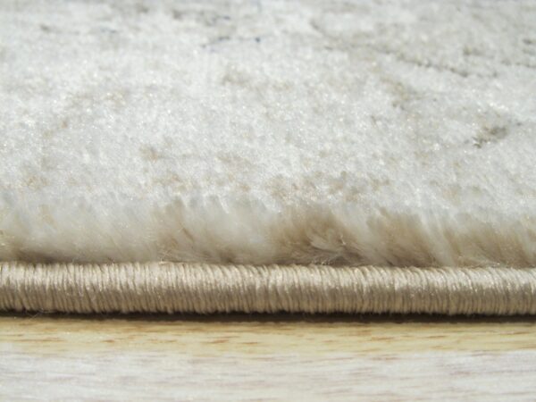 High-pile cream rug edge detail, showing soft fibers, subtle pattern, and tight beige binding on wood.