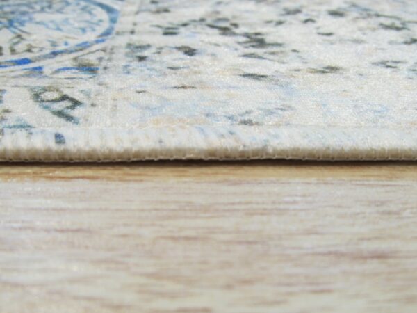 Close-up shows the thin, bound edge of a cream and blue rug against light wood flooring.