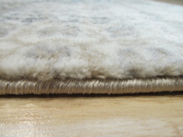 Close-up of the shaggy white pile rug edge with tan binding resting on a light wood floor.