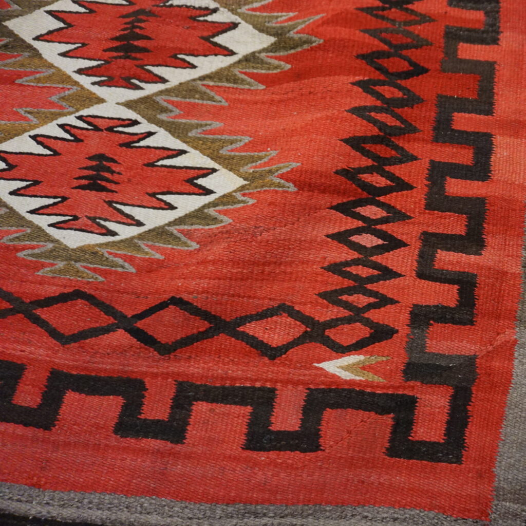 Close-up of a red flatweave rug featuring black, white, and brown zigzag diamond geometric patterns.