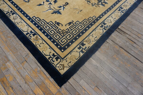 Tan rug with complex navy blue geometric and floral borders sits atop distressed gray wood flooring.