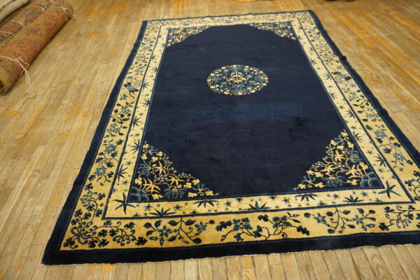 Blue rug with light floral border and medallion lays on a distressed wood floor near rolled carpets.