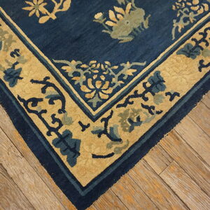Corner of a low-pile navy blue rug with gold and blue ornate borders on weathered wood flooring.