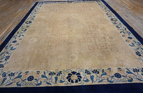 Tan rug featuring a subtle geometric field design and a prominent blue floral border on wood planks.
