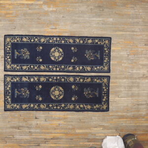 A pair of navy and gold patterned runners lie on a wood floor beside several rolled rugs.